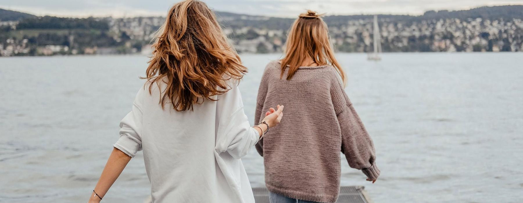 two women walk to the edge of a dock, in a body of water, looking toward the city on the other shore